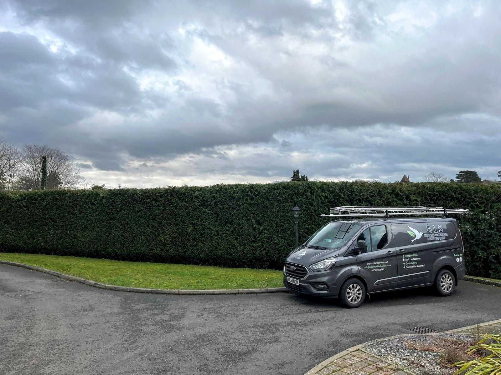 A BLS Ground & Garden van parked beside a long, neatly trimmed hedge.