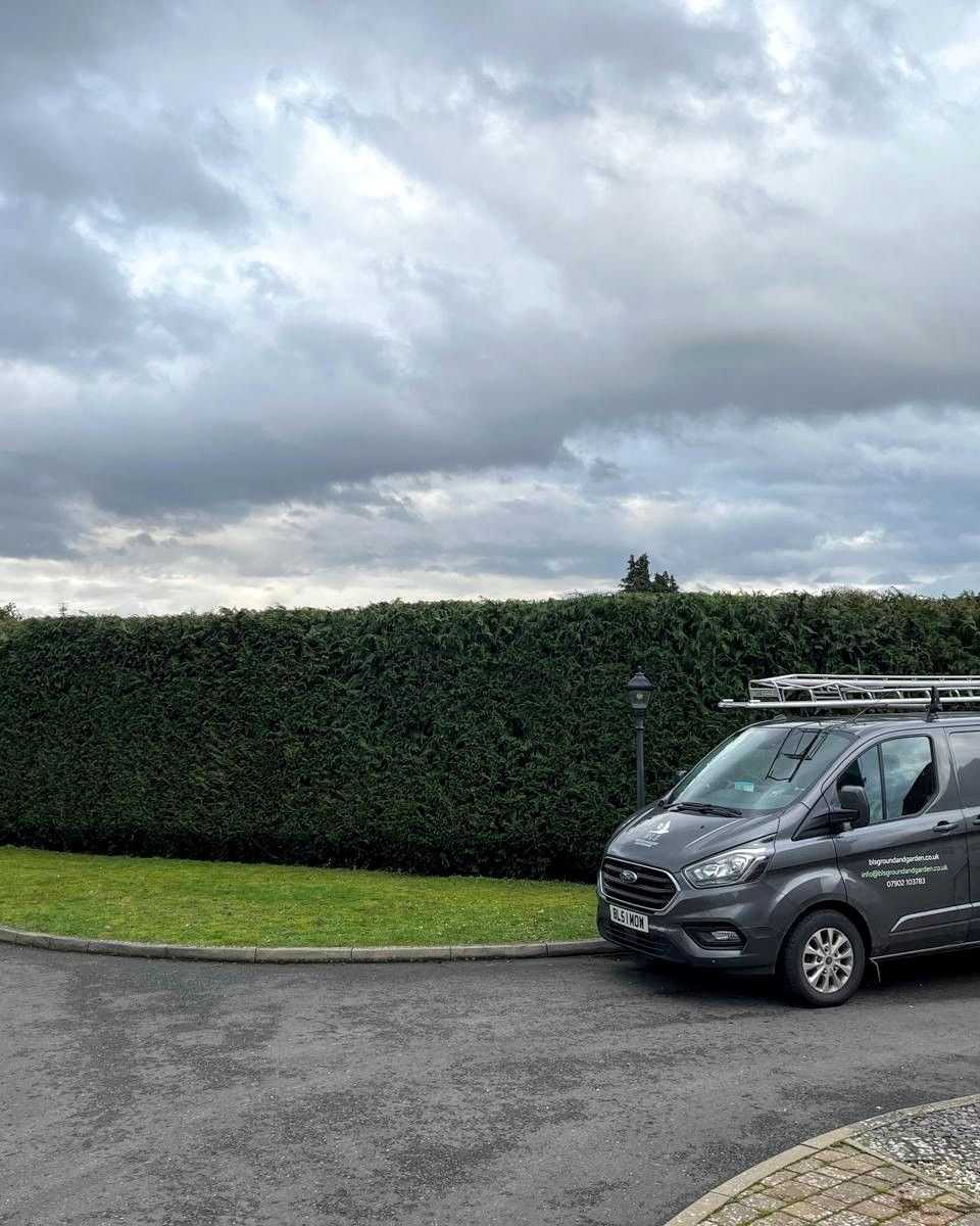 A BLS Ground & Garden van parked beside a long, neatly trimmed hedge.