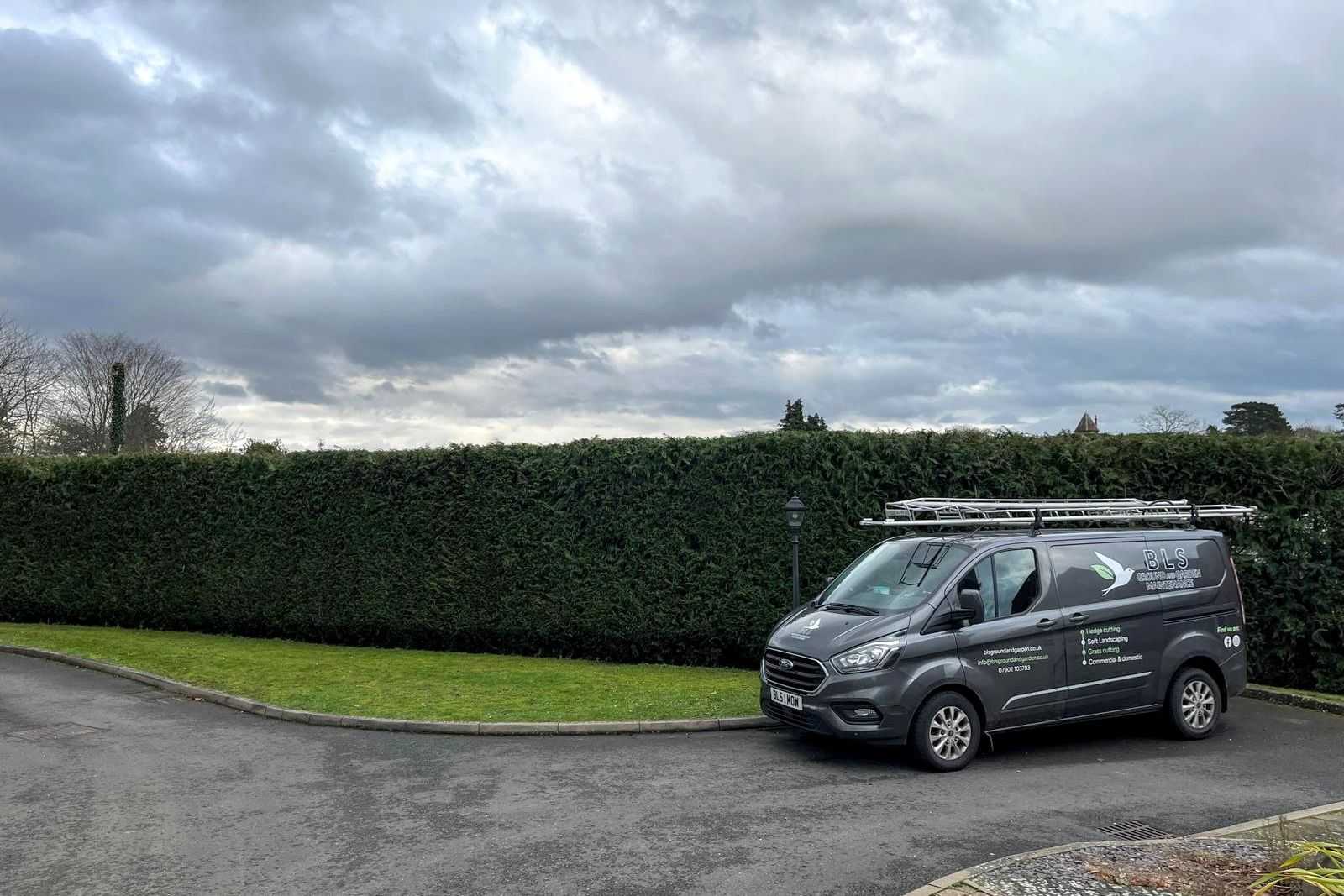 A BLS Ground & Garden van parked beside a long, neatly trimmed hedge.