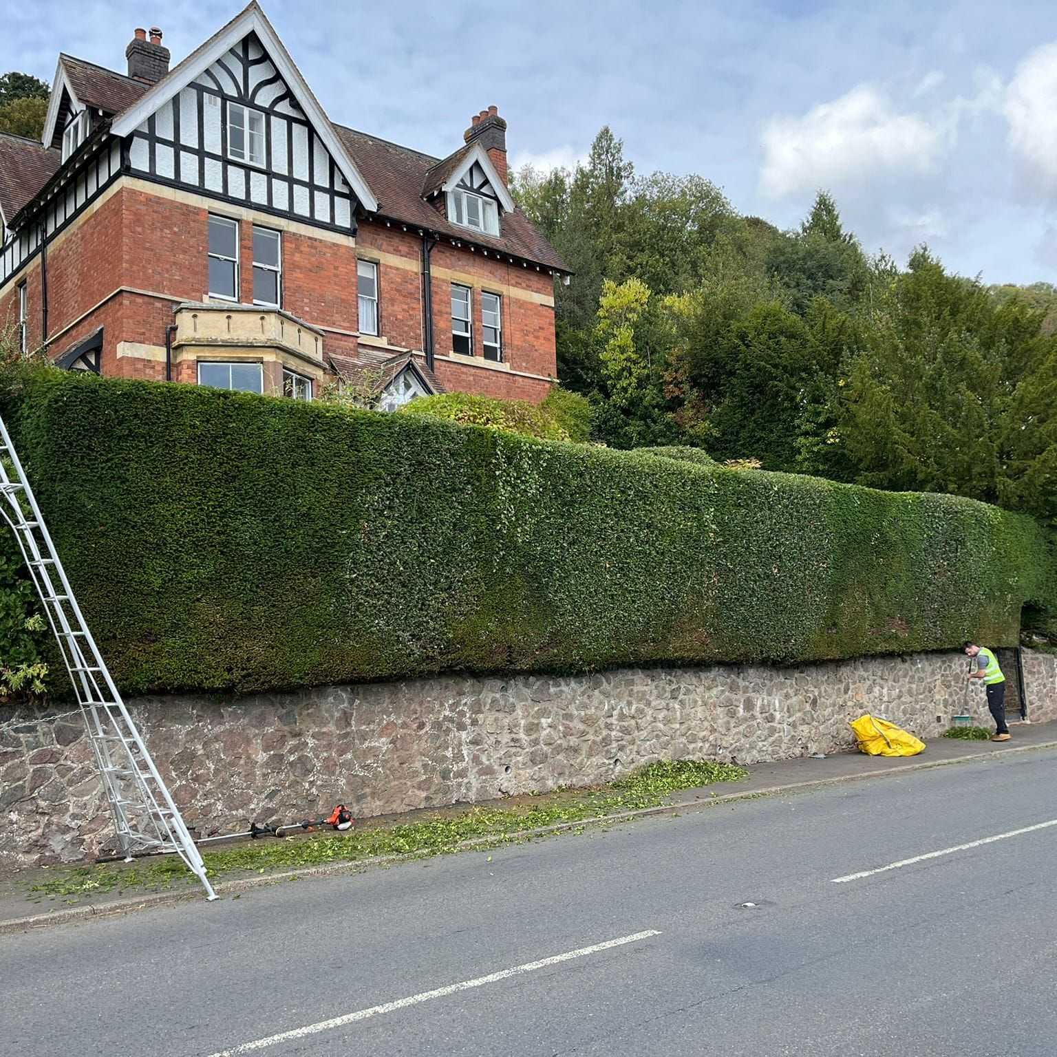 A long, neatly cut hedge along a roadside wall in front of a large red-brick house.