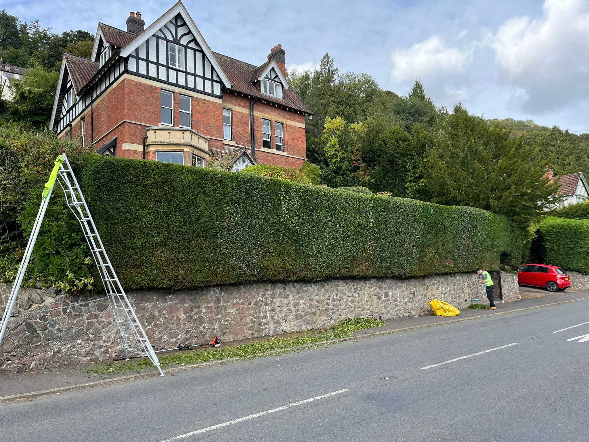 A long, neatly cut hedge along a roadside wall in front of a large red-brick house.